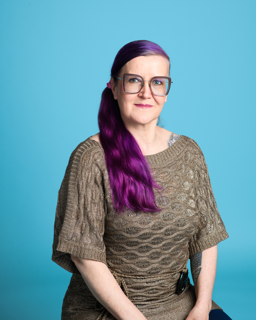 Mari istuu tasaisen vaalen sinist&auml; taustaa vasten jakkaralla ja hymyilee lempe&auml;sti pystypotrettikuvassa.