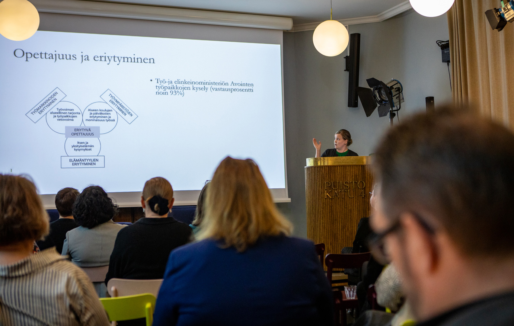 Kosunen esittelee tutkimustaan lavalla. Isolla kankaalla n&auml;kyy hieman vaikeasti tulkittava kuvio. Laaja kuva, jossa yleis&ouml;&auml; istuu selin kameraan.
