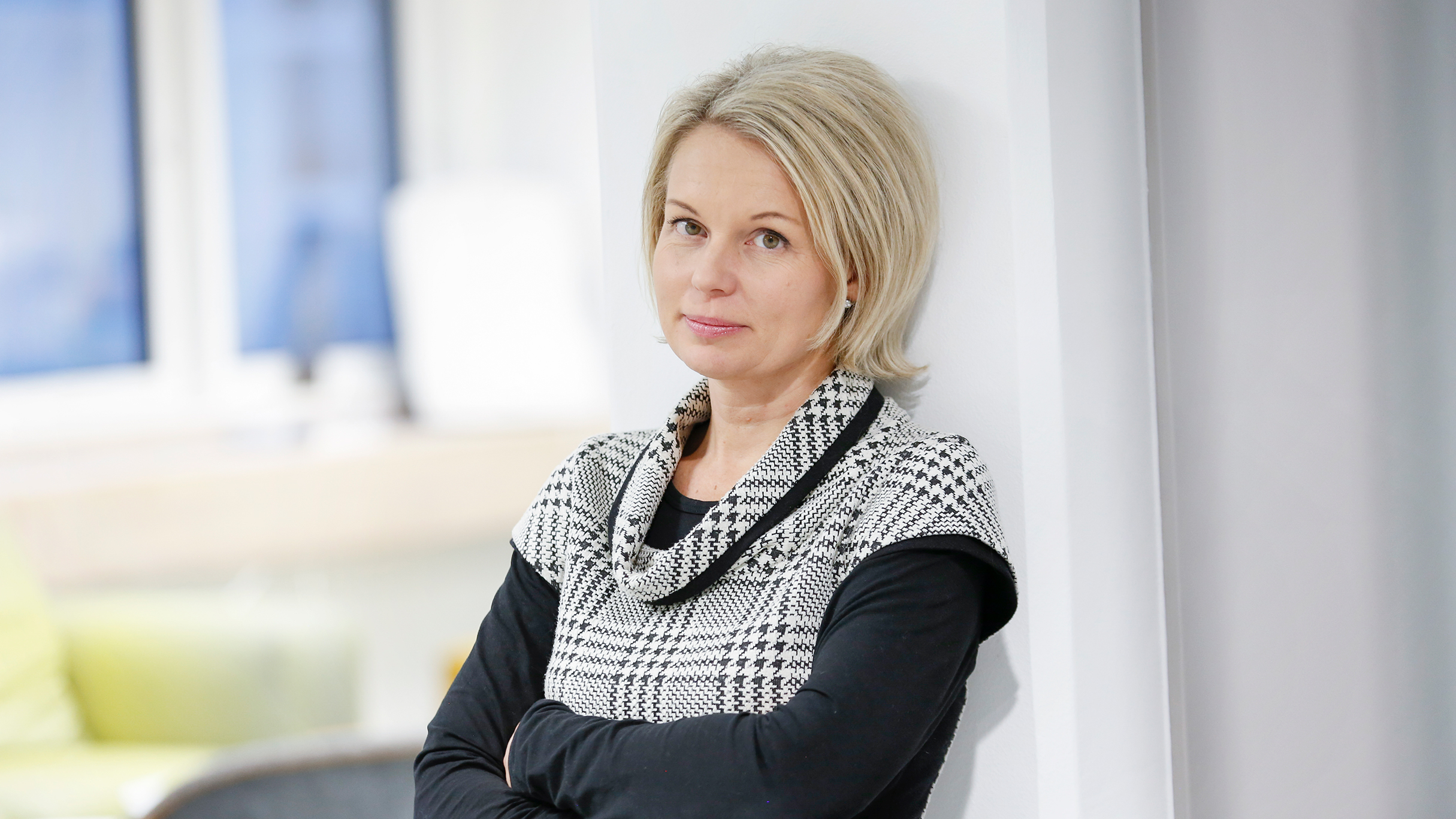 Nina Lahtinen katsoo kameraan kuvan keskellä yllään mustaharmaa asu. Taustalla vihreä sohva ja ikkuna. 