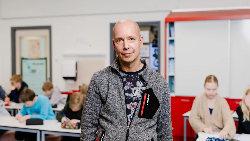 Mies harmaassa takissa katsoo kameraan kuvan keskellä. Hänen taustallaan on sumeana pöytiä, joiden ääressä on oppilaita työskentelemässä kirjojen äärellä.