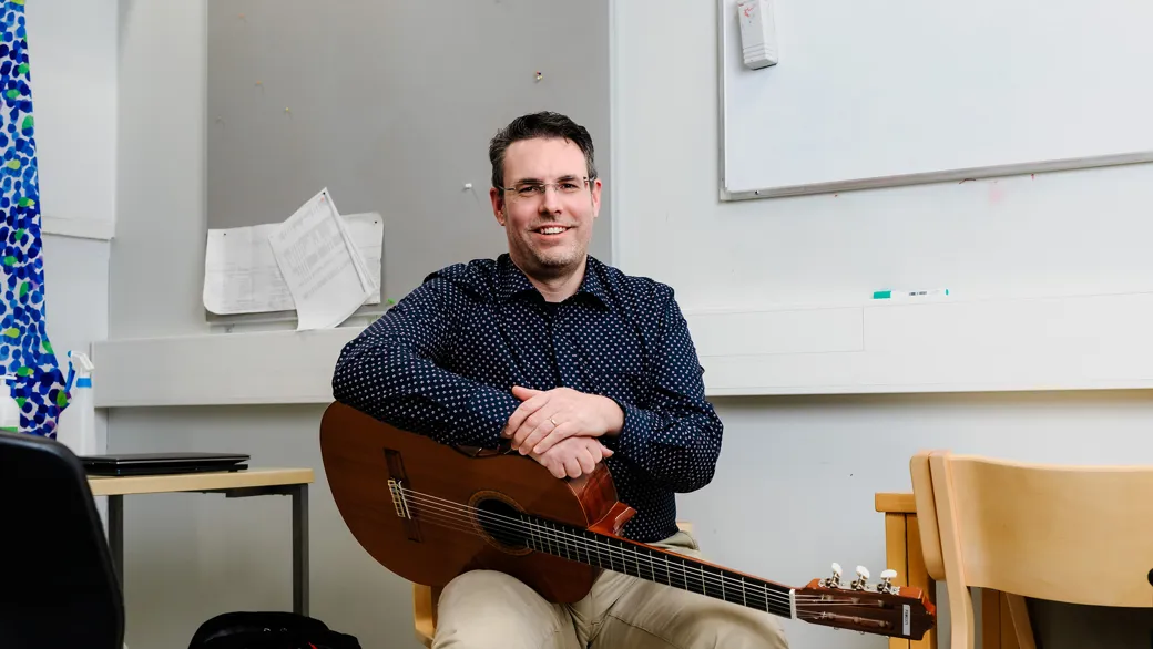 Mies istuu kitara sylissään ja katsoo kameraan. Hänellä on yllään tumma kauluspaita ja vaaleat housut. Taustalla näkyy seinä, jolla on harmaa ja valkoinen levy.