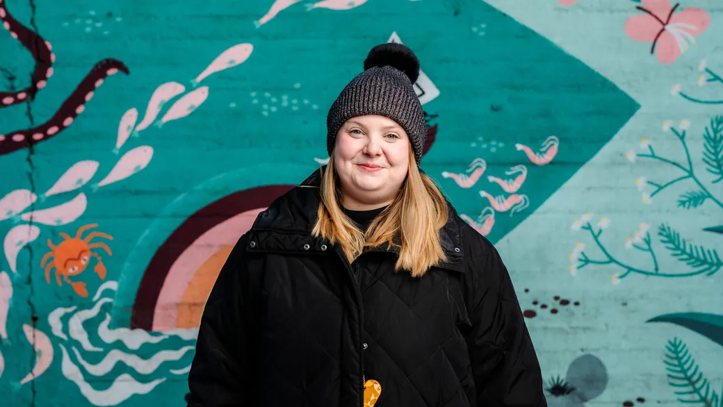 Roosa Tilles katsoo kameraan kuvan keskellä. Yllään hänellä on musta takki ja päässä pipo. Taustalla sinivihreän sävyinen seinä, johon on maalattu esimerkiksi kukkia.