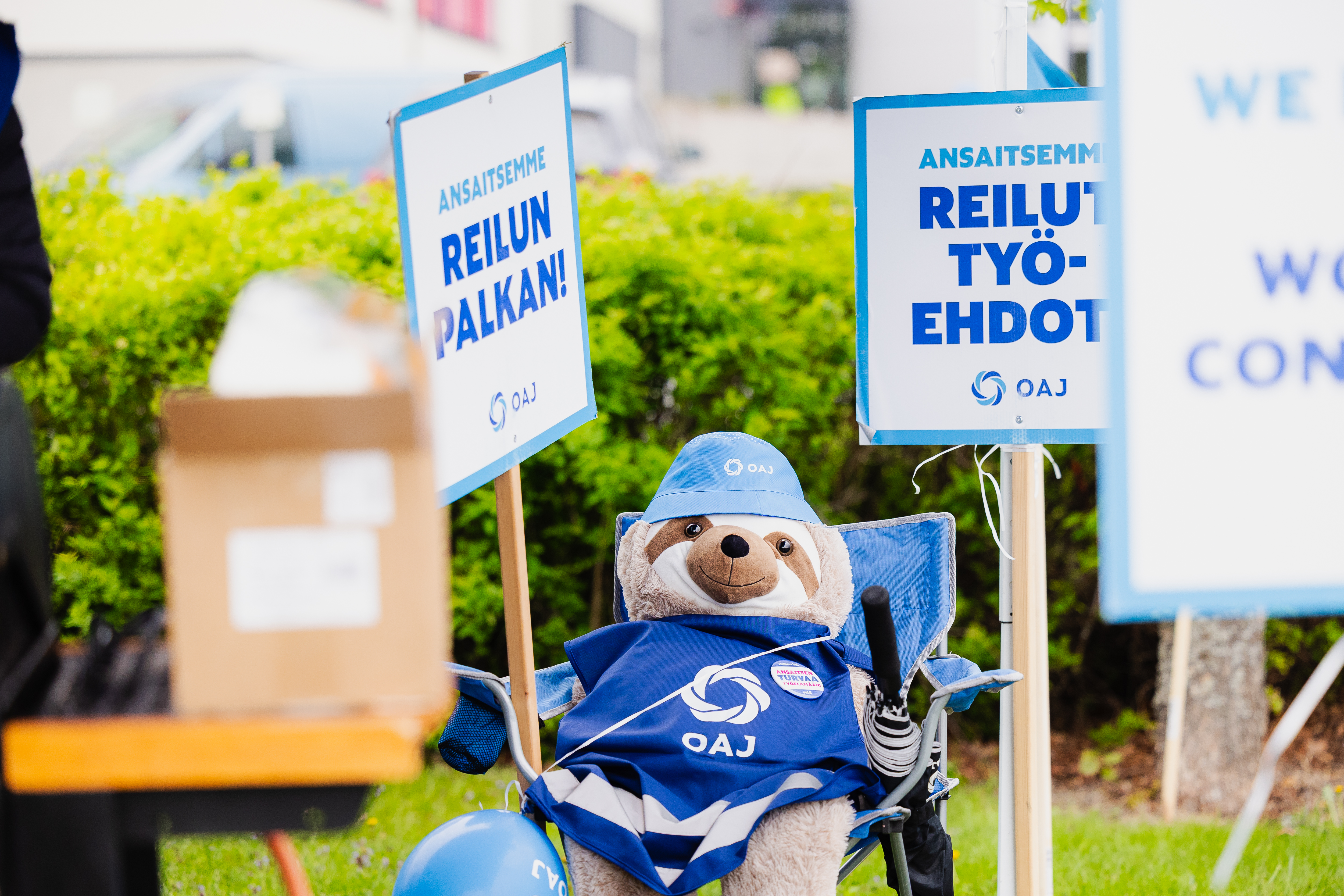 Kuvassa lakkokylttej&auml; sek&auml; OAJ:n lakkoliiviin puettu laiskiaispehmolelu, joka istuu retkituolilla.Kylteiss&auml; lukee "Ansaitsemme reilut ty&ouml;ehdot" ja "Ansaitsemme reilun palkan"
