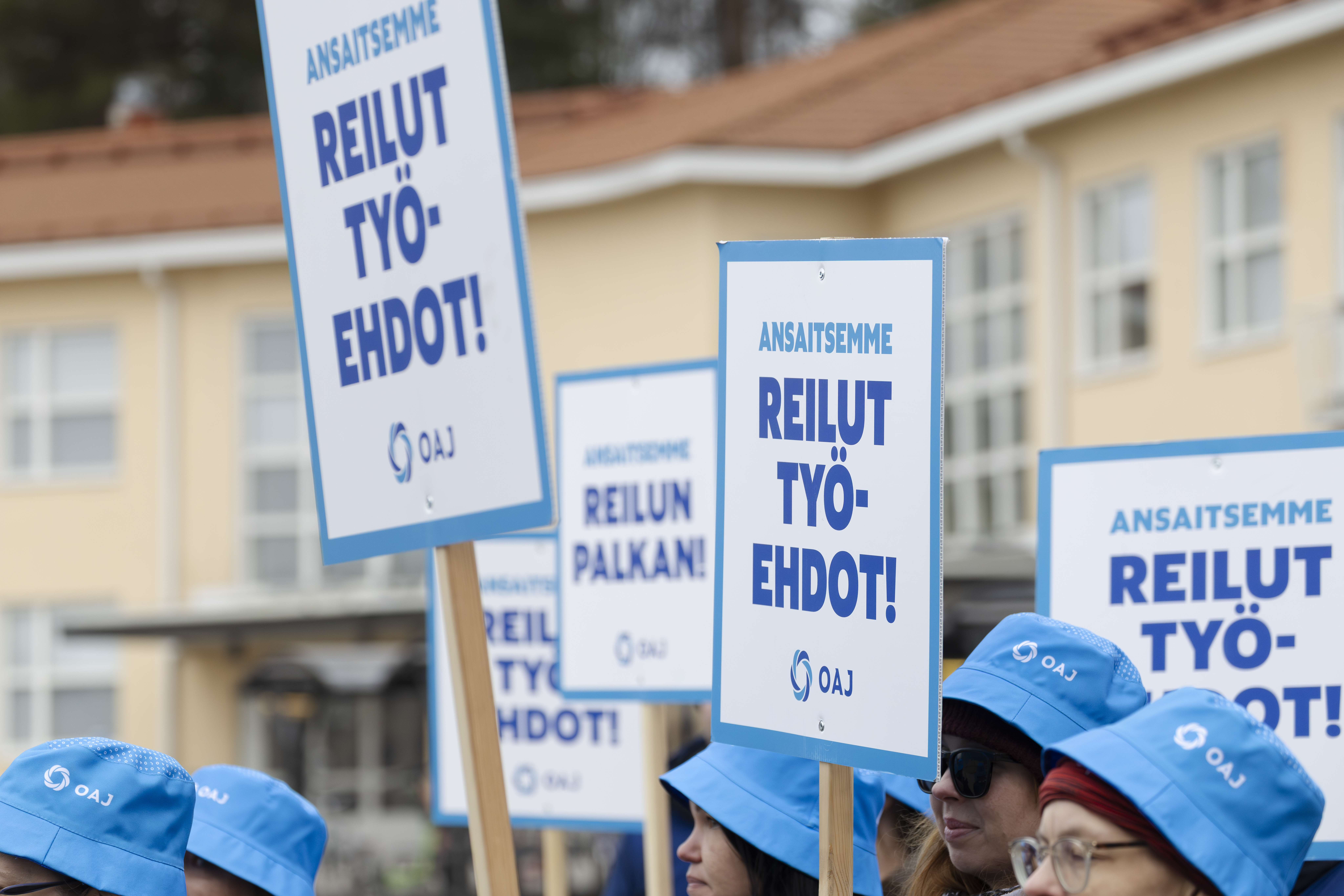 Lakossa olevia henkil&ouml;it&auml; seisoo kampuksen pihalla kyltit korkealla k&auml;siss&auml;&auml;n. Kuva on rajattu niin, ett&auml; lakkoilijoista n&auml;kyy vain kasvojen yl&auml;osa ja kyltit, joissa lukee "Ansaitsemme reilut ty&ouml;ehdot!" 