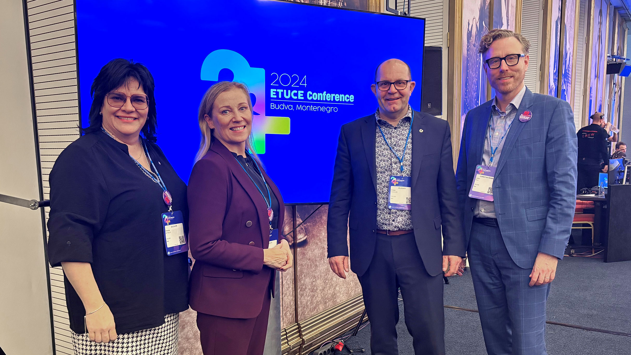 Neljä henkilöä seisoo näytön edessä, katsoo kameraan ja hymyilee. Näytöllä lukee: ETUCE Conference 2024 Budva, Montenergo.
