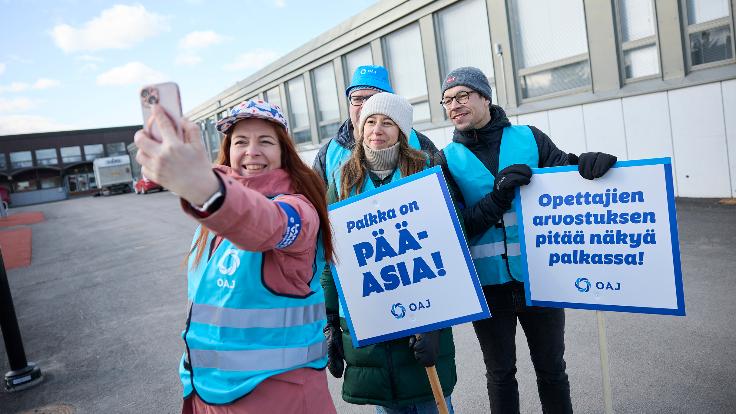 Neljä ihmistä seisoo siniset OAJ:n lakkovahti-liivit päällä koulurakennuksen edessä. Etummainen ottaa selfietä koko porukasta. Lakkovahdeilla on kyltit, joissa lukee Palkka on pääasia ja Opettajien arvostuksen pitää näkyä palkassa.