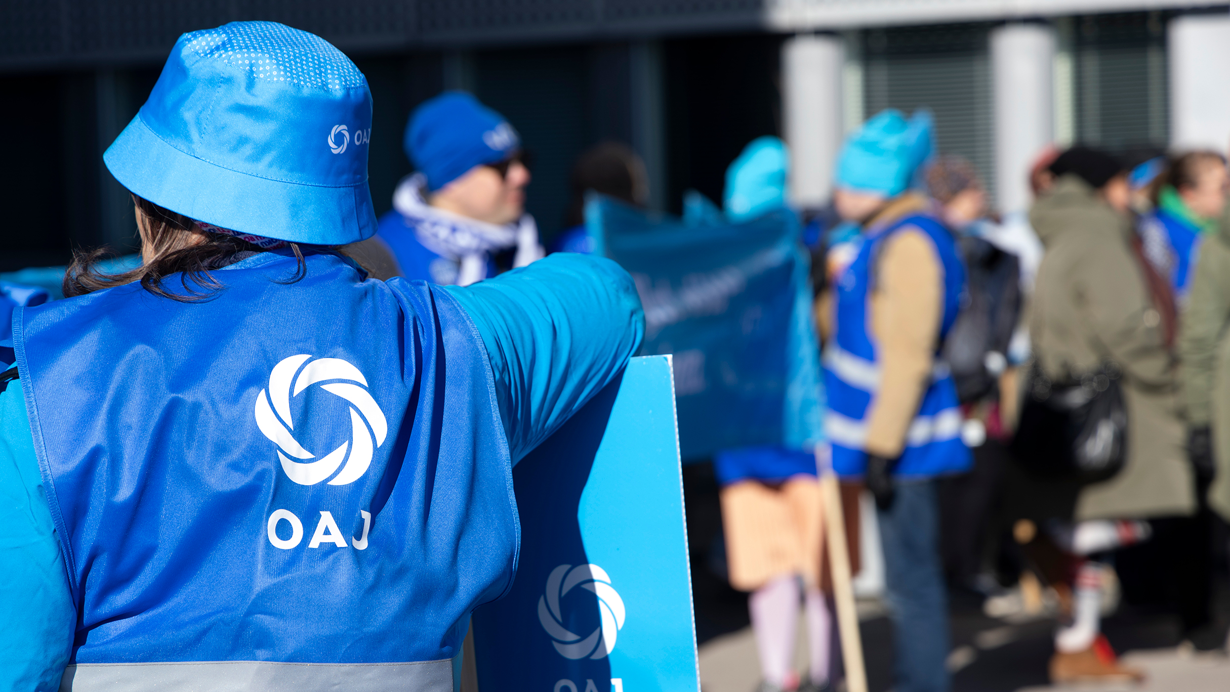 Ihminen selin kameraan, hänellä on sininen heijastinliivi, sininen kyltti ja sininen hattu. Kaikissa valkoiset OAJ-logot.